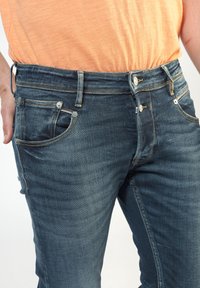 Jeans de denim azul oscuro con un corte slim, que presentan cinco bolsillos, costuras en contraste y herrajes en tono plateado en la parte frontal.