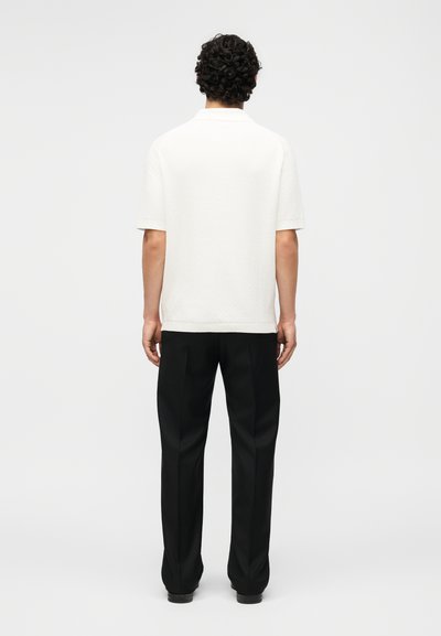 Άνδρας με σγουρά σκούρα μαλλιά στέκεται με την πλάτη, φοράει απλό λευκό κοντομάνικο πουκάμισο και μαύρο παντελόνι με ίσια ποδῶνα.