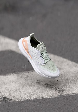 Jasnozielony i biały but biegowy Adidas z brzoskwiniowymi akcentami, zawieszony nad asfaltem z białymi oznaczeniami drogowymi.