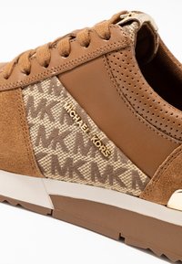 Sneaker in suede e pelle marrone con logo e dettagli dorati, dotata di un pannello in tessuto pattern che mostra "MK" in marrone chiaro su beige.