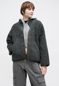 Γκρι σακάκι sherpa με κλείσιμο με κουμπιά, δύο μπροστινές τσέπες και άνετη εφαρμογή, φορεμένο πάνω από ένα ανοιχτό γκρι φούτερ με φερμουάρ.