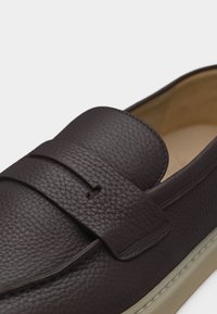 Σκούρο καφέ μοκασίνι από υφή δερμάτινη με λουράκι στο πάνω μέρος και εσωτερική επένδυση και σόλα σε μπεζ χρώμα και με μαξιλαρωτό πάτο.