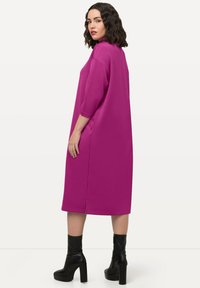 Fuchsiové oversize šaty s 3/4 rukávy a bočními kapsami, s vysokým výstřihem a rozparkem na lemu, doplněné černými botami na podpatku.