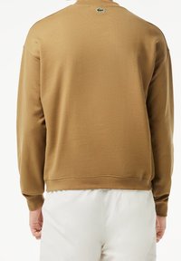 Hnědá mikina s měkkou texturou, žebrovanými manžetami a spodním lemem. Logo Lacoste v zelené barvě na zadním výstřihu, doplněné bílými kalhotami.