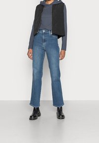 Black quilted vest over a blue hoodie, paired with high-waisted denim flared jeans and black ankle boots. Smooth textures, simple design.