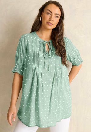 Femme portant une blouse ample vert menthe avec des pois rectangulaires blancs, des détails plis couchés, un col à nouer et des manches courtes élastiquées froncées.