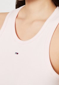 Detailní záběr na osobu oblečenou v svetle růžovém žebrovaném tílku s malou obdélníkovou červenou, bílou a modrou nášivkou na hrudi.