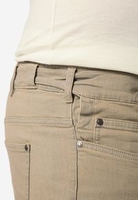 Beige farkut sileällä pinnalla, joissa on viisi taskua, vyölenkit ja metallikoristeet. Lähikuva sivusta ja vyötäröltä.