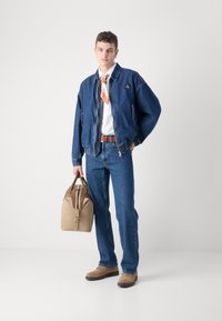 Veste bomber en denim, bleue, fermeture éclair, logo sur la poitrine. Chemise blanche, ceinture marron, sac beige, jeans bleus, chaussures beiges. Textures lisses.