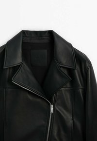 Veste en cuir noire avec un large col et une fermeture éclair argentée asymétrique partiellement zippée sur fond blanc.