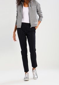 Šedá mikina na zip s logem Calvin Klein, bílý top na ramínka, černé pruhované kalhoty a stříbrné sportovní boty.