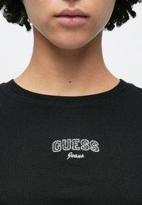 Czarna prążkowana koszulka z haftowanym białym napisem „GUESS Jeans” w kształcie łuku. Dopasowany dekolt z gładkiej, teksturowanej tkaniny.