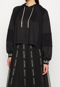 Czarna krótka bluza z kapturem, ozdobiona błyszczącymi srebrnymi sznurkami i kontrastowymi panelami. Noszona na czarnej siateczkowej spódnicy z tekstowymi akcentami.