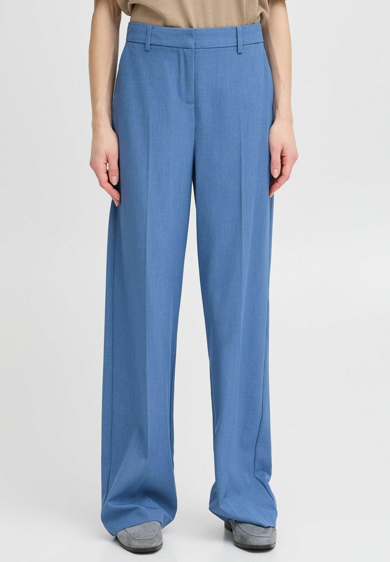Personne portant un pantalon bleu ample taille haute avec des plis devant et des chaussures grises, debout devant un fond clair uni.