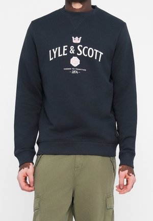 Persona que lleva una sudadera negra de Lyle & Scott con logo blanco y corona, combinada con pantalones cargo verde oliva, de pie frente a un fondo sencillo.