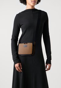 Brązowa, teksturowana torba na ramię z czarnym paskiem. Ma prostokątny kształt, zapięcie na zamek błyskawiczny oraz metalowe logo na przodzie.