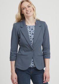 Tume sinine bleiser, millel on sälguga krae, üks nupp sulgemiseks ja kaks esimest taskut. Kandma mustrilise sinise pluusi peal. Löargs sobivus.