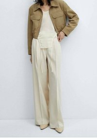 Braune kurz geschnittene Jacke mit zwei vorderen Taschen, cremefarbenes Oberteil und lockere, cremefarbene Hose mit Kordelzug. Beigefarbene spitze Schuhe vervollständigen das Outfit.