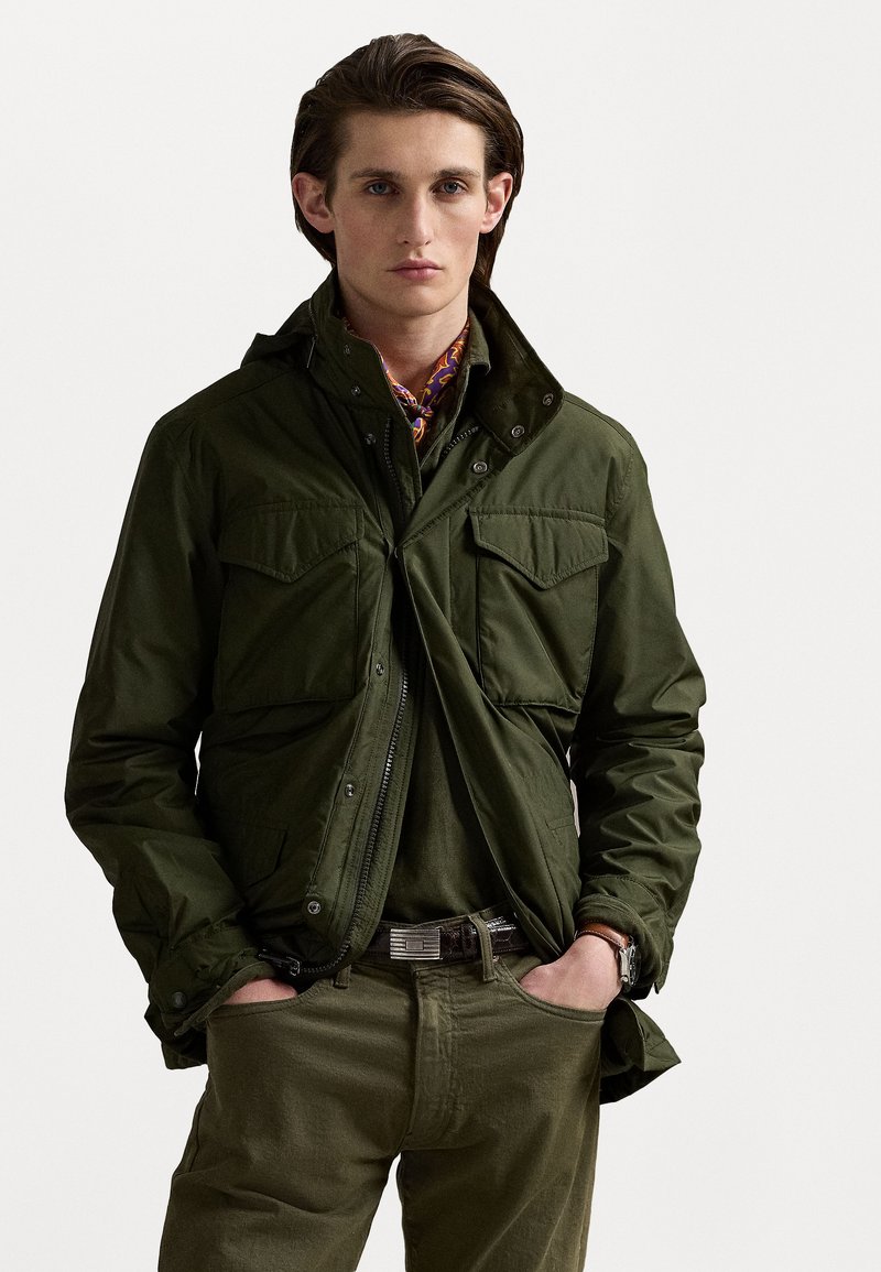 Olivgrüne Jacke mit Reißverschluss vorne, zwei Brusttaschen und Kapuze. Getragen über einem passenden Hemd, mit einem gemusterten Schal und Gürtel.