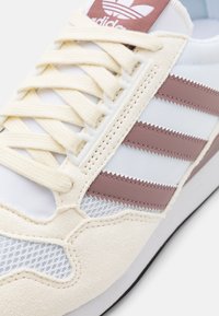 White athletic shoe with pink and cream accents, featuring three pink leather stripes, textured mesh panels, and flat cream laces.