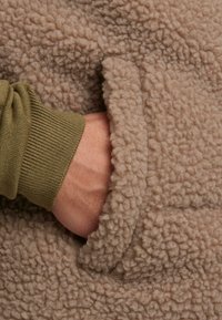 Barna bolyhos anyag, amelynek puha, texturált felülete van. Egy olívazöld mandzsettás kéz nyúl a side zsebbe, bemutatva a ruha kialakítását.