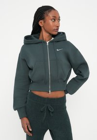Tamnozeleni skraćeni dukserica s prednjim patentnim zatvaračem, podešivim šnalama i kenguruvim džepom; sadrži Nike logo u bijeloj boji. Mekana tekstura tkanine.