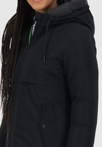 Giacca piumino nera con cappuccio, dotata di zip frontale, coulisse regolabile e tasca laterale con bottone a scatto. Texture liscia.