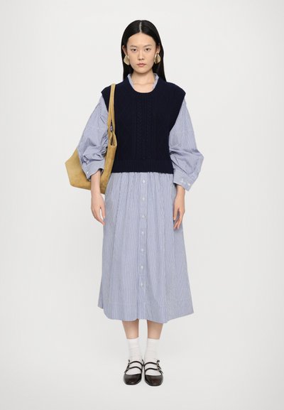 Gestreiftes blau-weißes Hemdkleid, geschichtet mit einem marineblauen Strickweste. Accessoires umfassen große, runde Ohrringe und eine beige Schultertasche.