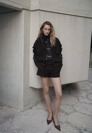 Braune Wildlederjacke und Shorts, kombiniert mit einem grau gestreiften Hemd. Dazu spitze braune Absätze. Das Modell posiert vor einer strukturierten Wand.