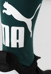 Zielona bawełniana skarpetka z dużym białym logo "PUMA" i sylwetką skaczącego kota. Czarny but z widocznym teksturowanym materiałem w okolicy kostki.