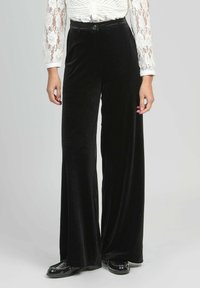 Pantalons larges en velours noir avec une texture lisse, taille haute et fermeture à bouton. Associés à un haut en dentelle blanche transparente.