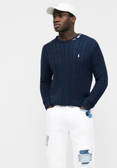 Άνδρας που φοράει μπλε πουλόβερ με πλέξη καναλιού, λευκό τζιν με φθορές και μπλε patches, και λευκό καπέλο μπέιζμπολ, στέκεται με τα χέρια στις τσέπες.