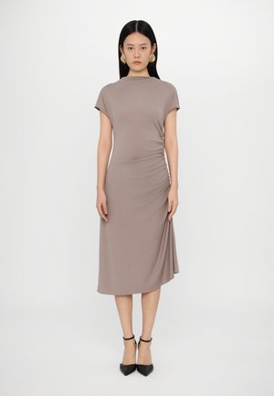 Robe mauve de longueur genou avec des manches courtes, présentant un détail de fronce sur le côté. Portée avec des escarpins noirs à talons pointus et des boucles d'oreilles sculpturales dorées.