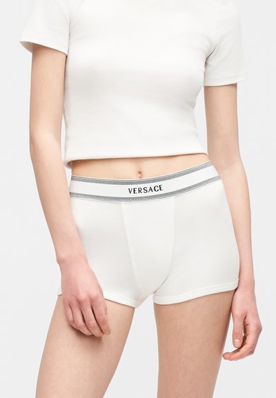 Γυναίκα που φοράει λευκό crop top με κοντά μανίκια και λευκά boxer με το λογότυπο «VERSACE» στη μέση, στέκεται μπροστά σε λευκό φόντο.