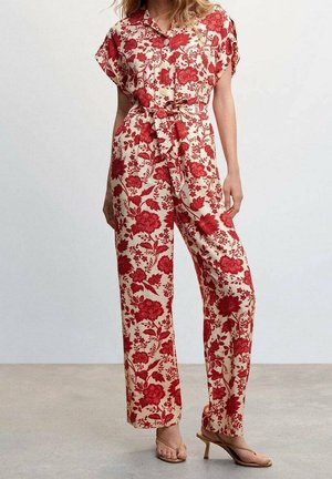 Vrouw draagt een beige overall met rode bloemenprint, korte mouwen, een tailleband en beige haksandalen tegen een effen achtergrond.