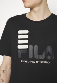 T-shirt in cotone nero con un grande logo "FILA" stilizzato in grigio e bianco, con dettagli a strisce e scollatura rotonda.
