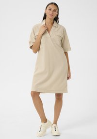 Beige randig skjortklänning med krage, korta ärmar och två bröstfickor. Bärs med kraftiga vita sneakers. Slät tygtextur.