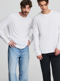 Białe, bawełniane koszulki z długim rękawem i okrągłym dekoltem. Jedna w połączeniu z jasnoniebieskimi dżinsami, druga z czarnymi spodniami.