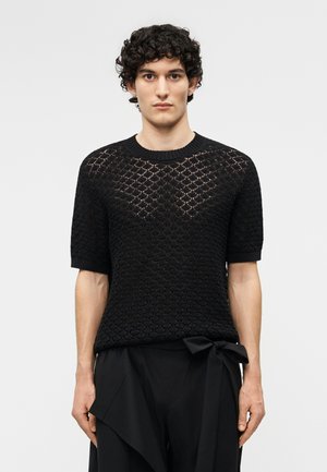 Jeune personne aux cheveux bouclés portant un haut en maille à manches courtes noir à motifs et un pantalon noir avec un nœud noué à la taille.