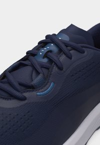 Κοντινό πλάνο ναυτικού μπλε αθλητικού παπουτσιού με υφασμάτινη υφή, επίπεδα κορδόνια και μπλε δερμάτινη ετικέτα κορδονιών που εμφανίζει μερικό λευκό κείμενο.