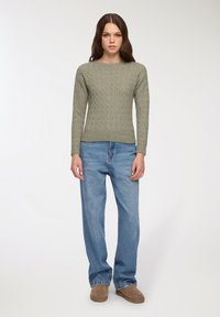 Kobieta stoi w zielonym swetrze w warkocze, luźnych niebieskich dżinsach i brązowych zamszowych butach na tle jednorodnym jasnym.
