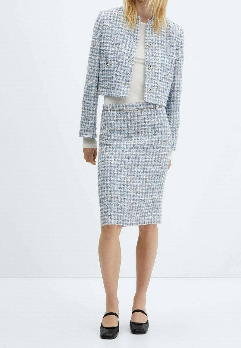 Femme portant une veste courte en tweed bleu clair et blanc assortie à une jupe crayon mi-longue, avec des chaussures Mary Jane noires.