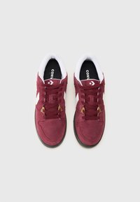 Burgundy suede sneakers with white accents, featuring black insoles, flat laces, and a low-top design. Rubber sole with a textured finish.