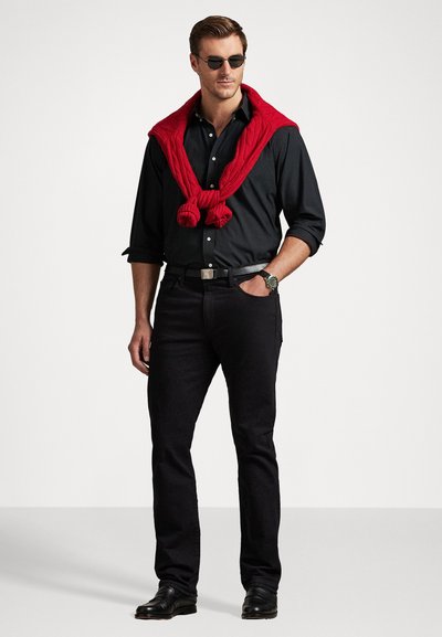 Svart knappskjorte, svarte jeans og svarte sko. En rød genser ligger over nakken. Tilbehør inkluderer mørke solbriller og et armbåndsur.