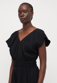 Black dress with a V-neck, short sleeves, and gathered waistband. Made of textured fabric, it falls loosely to the waist.