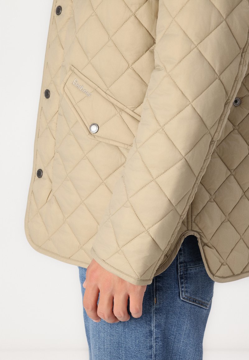 Beige gesteppte Jacke mit einem Diamantmuster, ausgestattet mit einem Druckknopfverschluss und einer Seitentasche. Weiche Textur, kombiniert mit blauen Jeans.