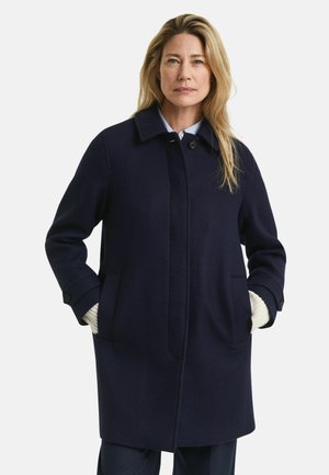 Navy wool coat with a straight cut, buttoned front, and two large pockets. Features a rounded collar and ribbed sweater cuffs peeking out.