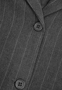 Szary żakiet w prążki z dwoma czarnymi guzikami z czterema otworami. Tkanina ma fakturowe wykończenie, podkreślające pionowe paski.