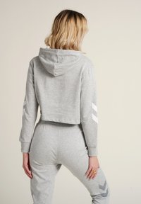 Hoodie gris court avec capuche, présentant des accents en chevron blancs sur les manches. Tissu doux avec une texture lisse, associé à un jogging assorti.