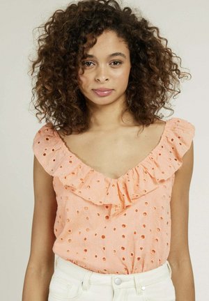 Blouse pêche avec un col à volants, présentant des découpes en œillet, une texture douce et une coupe décontractée, associée à un jean de couleur claire.